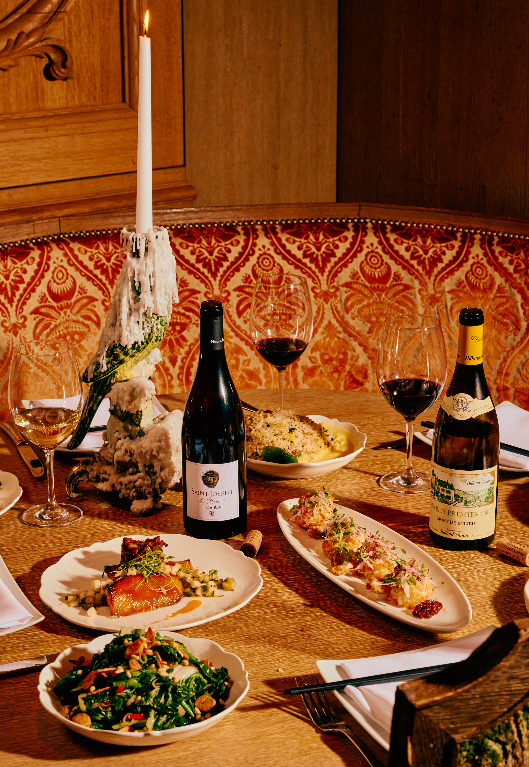 A restaurant table set with multiple dishes, two wine bottles, red and white wine glasses, and a lit taper candle in front of a patterned red booth.