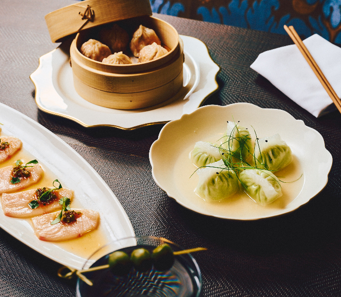Three dim sum dishes on a table including a bamboo steamer with dumplings, jade-colored dumplings in broth, and sliced fish with garnish.