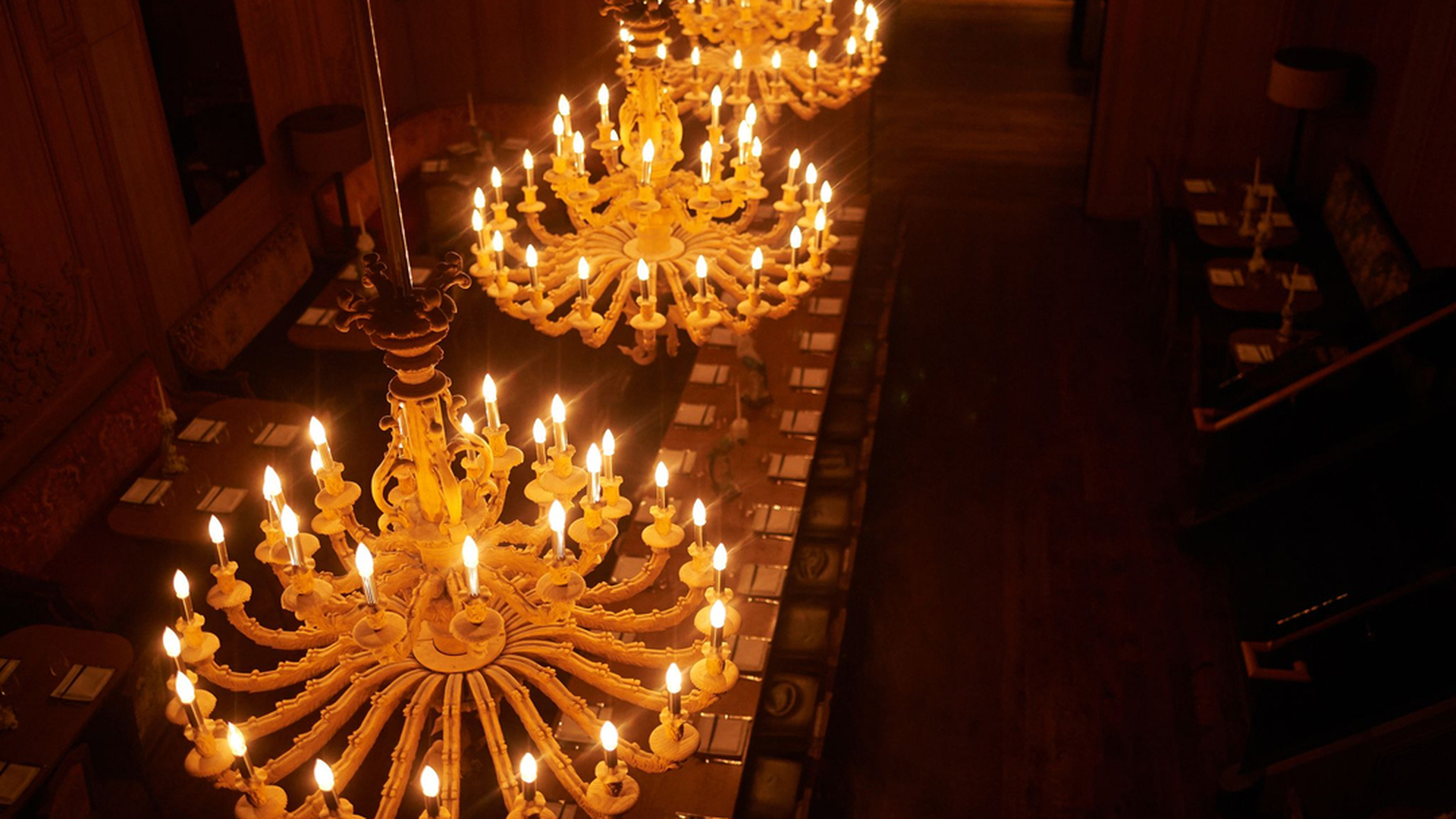 A long, ornate chandelier with many lit candles hangs above a wooden floor, casting a warm glow.