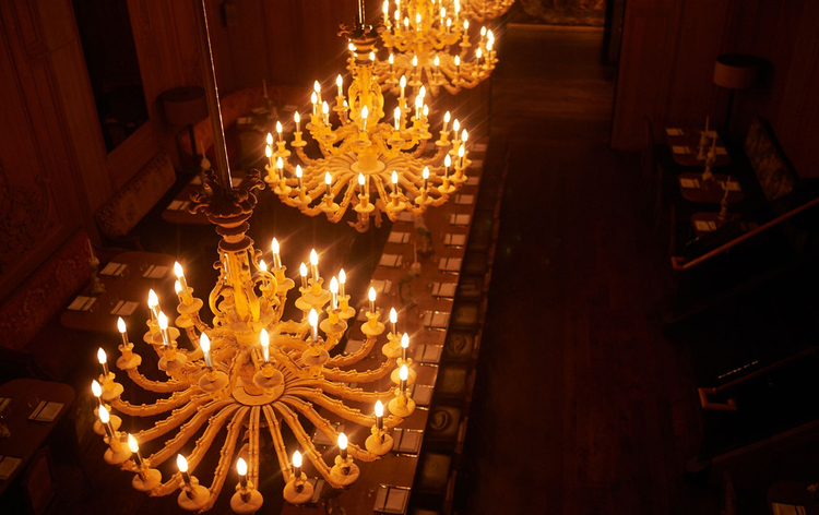 Grand chandeliers with numerous lit candles hang above a long table in a dimly lit room, suggesting a formal dining atmosphere.