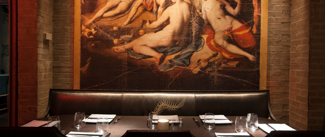 A restaurant booth with a dark table setup against a brick wall, featuring a large painting of nude figures and musical instruments above.